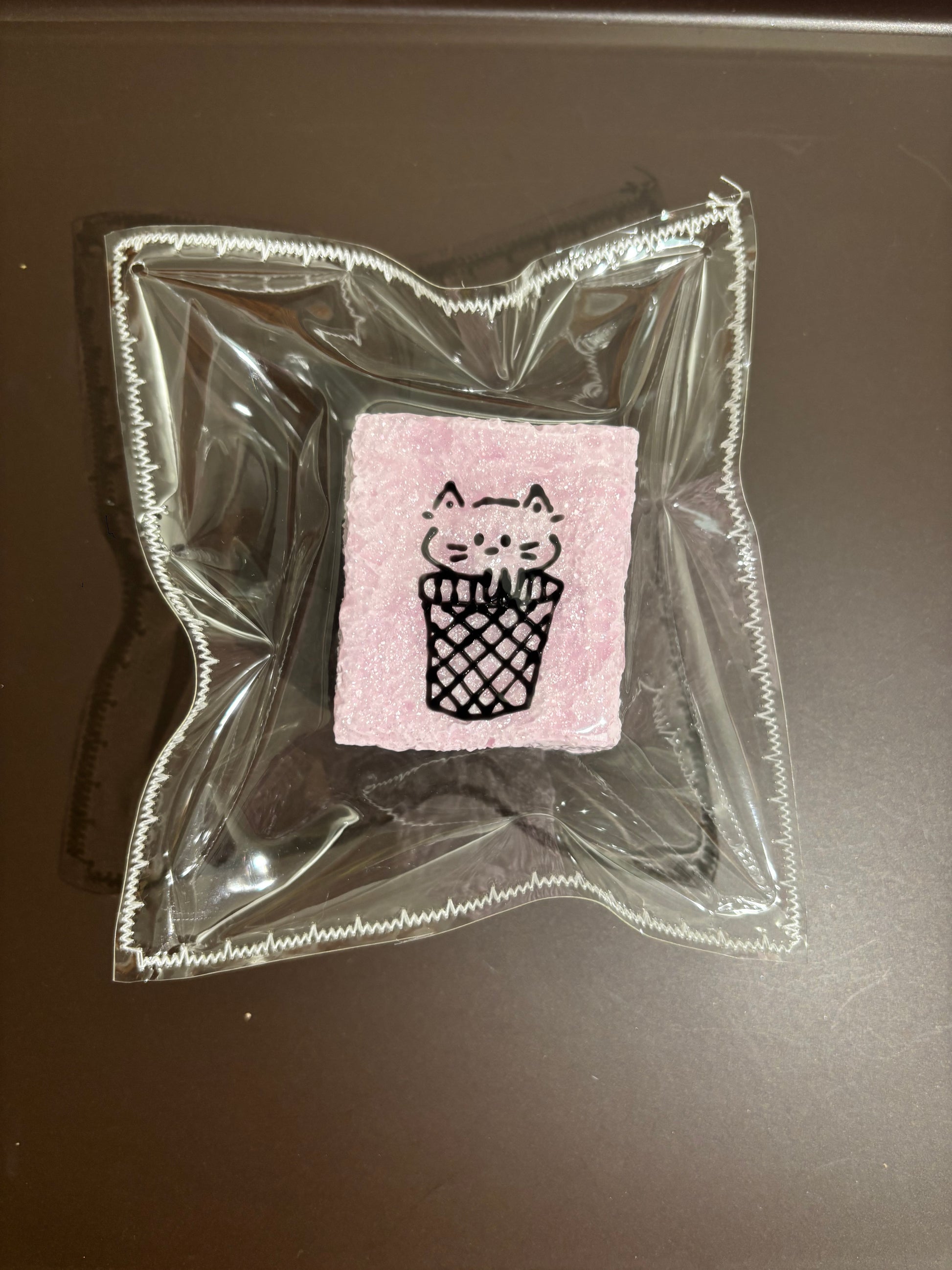 透明袋入りの白い刺繍布。口元があるネコ風キャラとバスケット模様の刺繍。