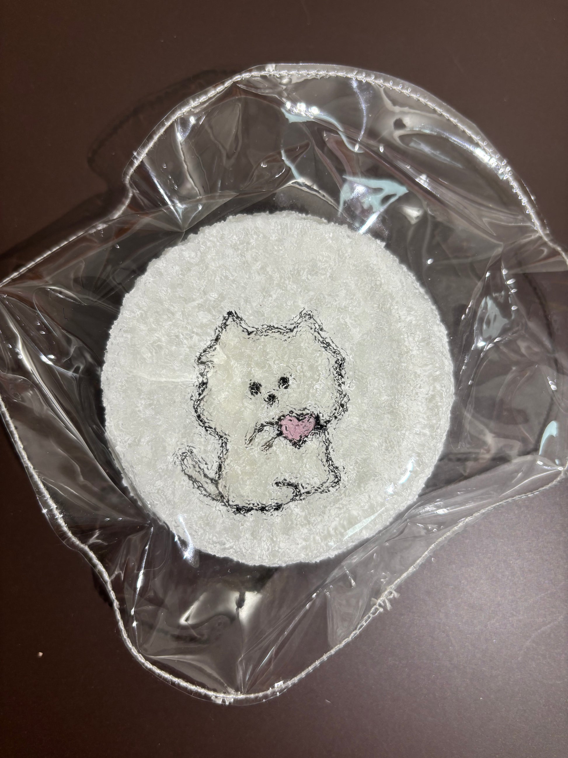 透明袋入りの白い刺繍布。丸いクマ風顔刺繍、中心にピンクの小さな差し色。