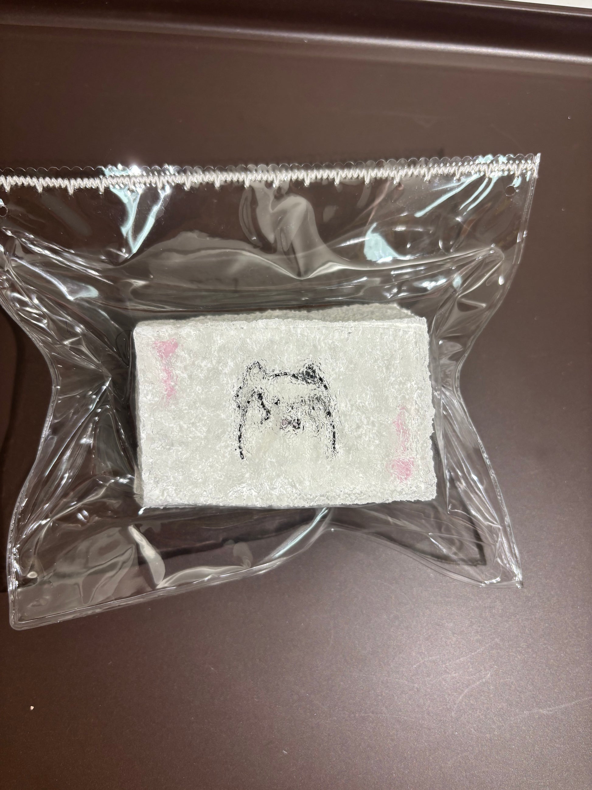 透明袋入りの白い刺繍布。クマ風の顔と小さな点々、ピンクの差し色。