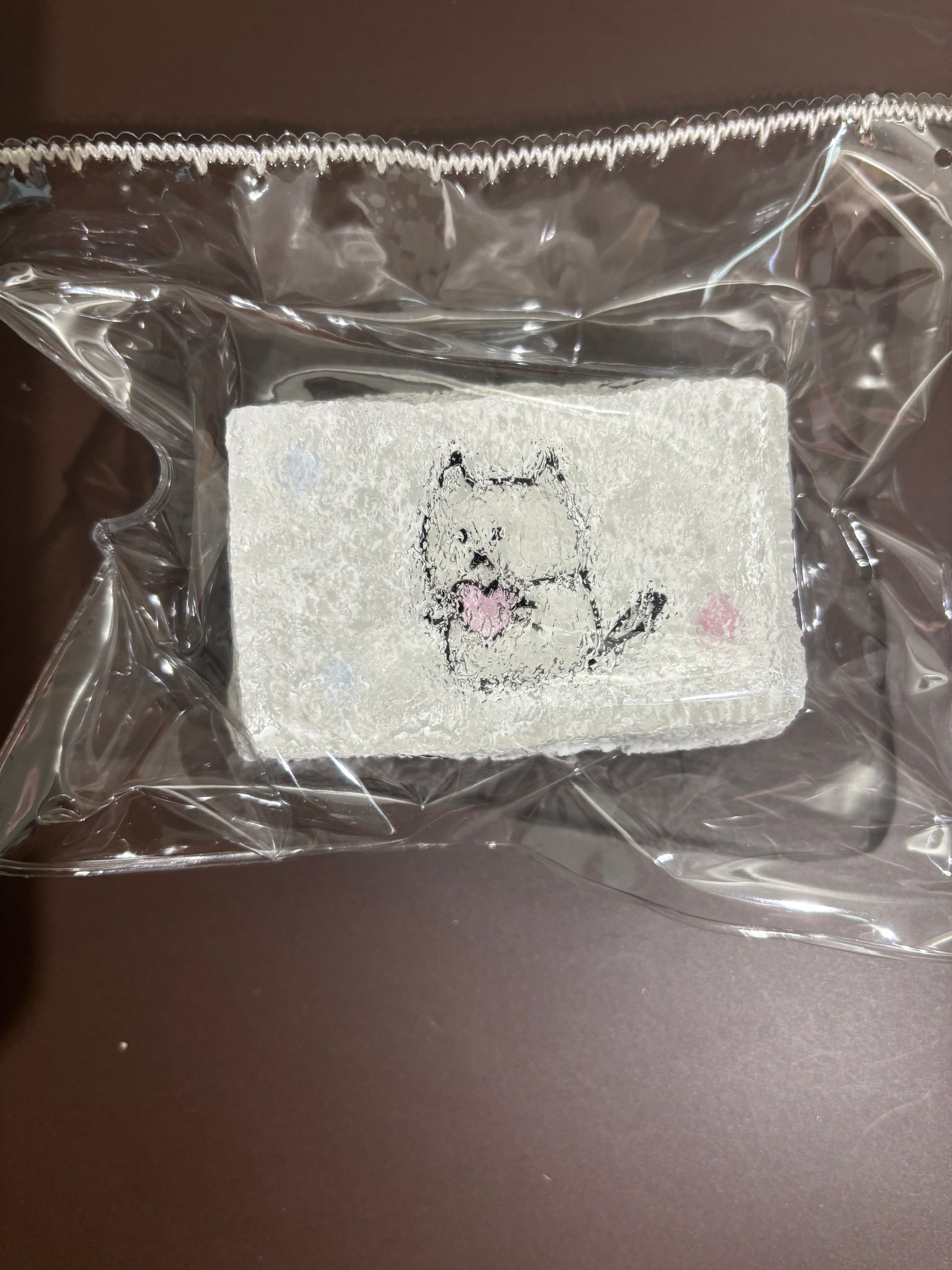透明袋入りの白い刺繍布。クマ風の顔の刺繍、全体に薄いピンクのアクセント。